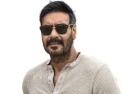 Ajay Devgn Stylish Man With Dark Hair, Beard, And Sunglasses, Wearing A Casual Beige Shirt, Against A White Background, Representing Celebrity Style Or Entertainment Industry.