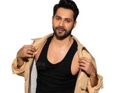 Varun Dhawan Stylish Man In Black Tank Top And Beige Jacket, Posing Confidently With Hand On Collar, Showcasing Fashion And Personality.